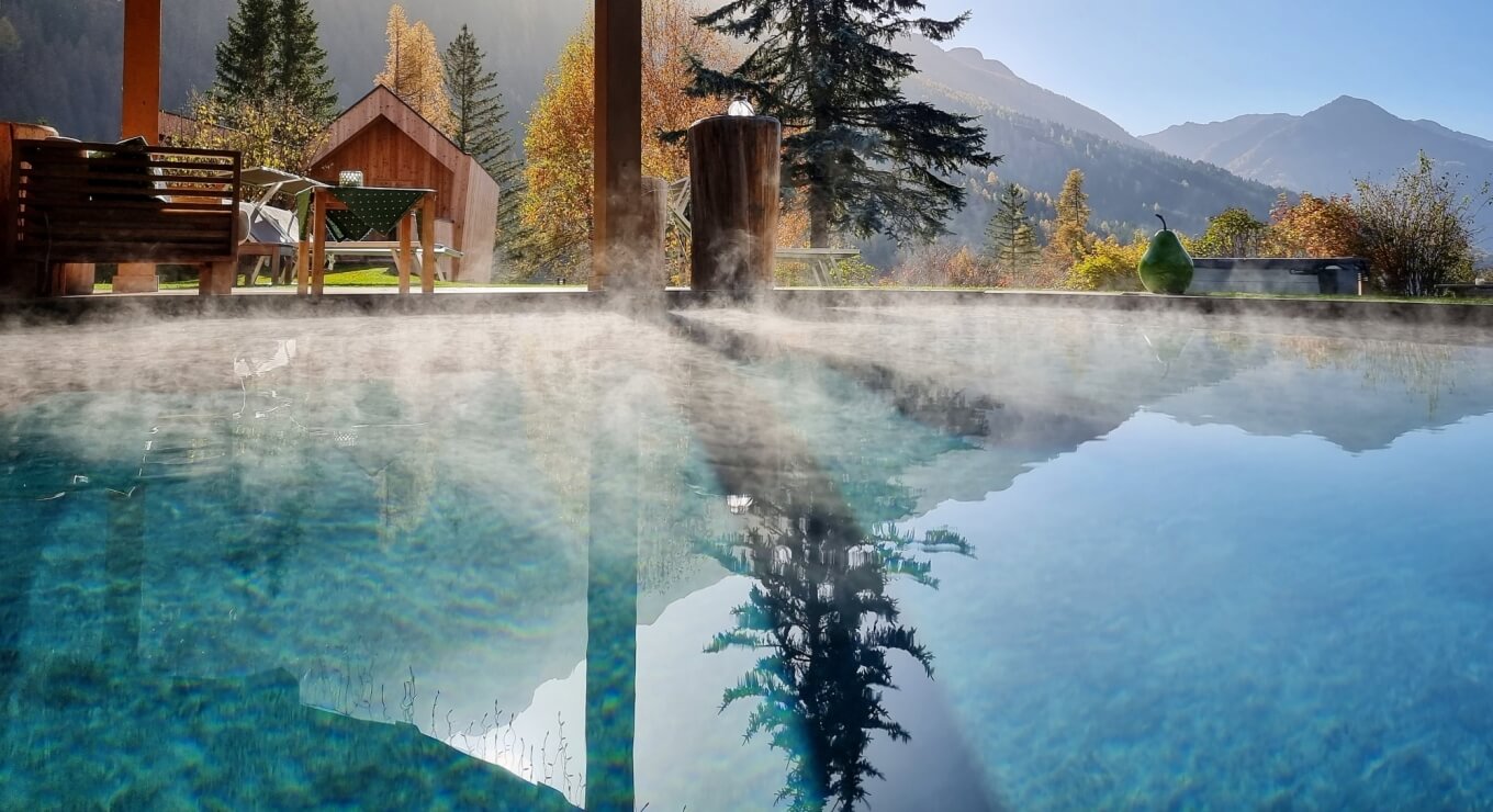 Una piscina all'aperto con acqua blu, montagne e alberi autunnali, una panchina e una scultura di pera verde.