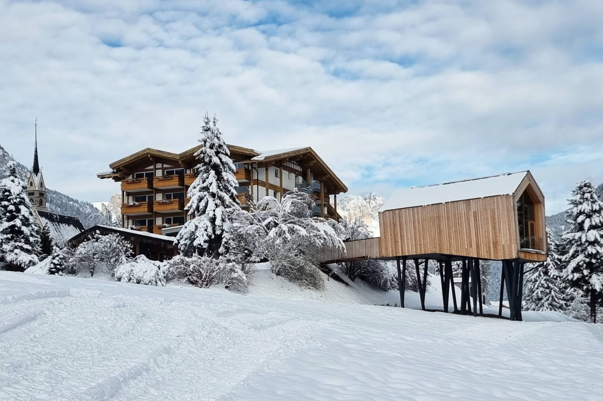 Paesaggio invernale sereno con una grande casa di legno e pietra e un piccolo edificio su palafitte, entrambi coperti di neve.