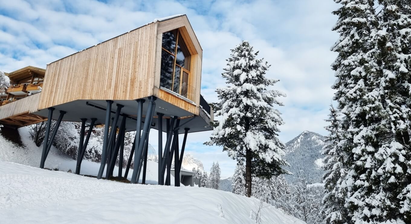 Struttura moderna in legno su palafitte, con finestra triangolare, circondata da alberi e montagne innevate.