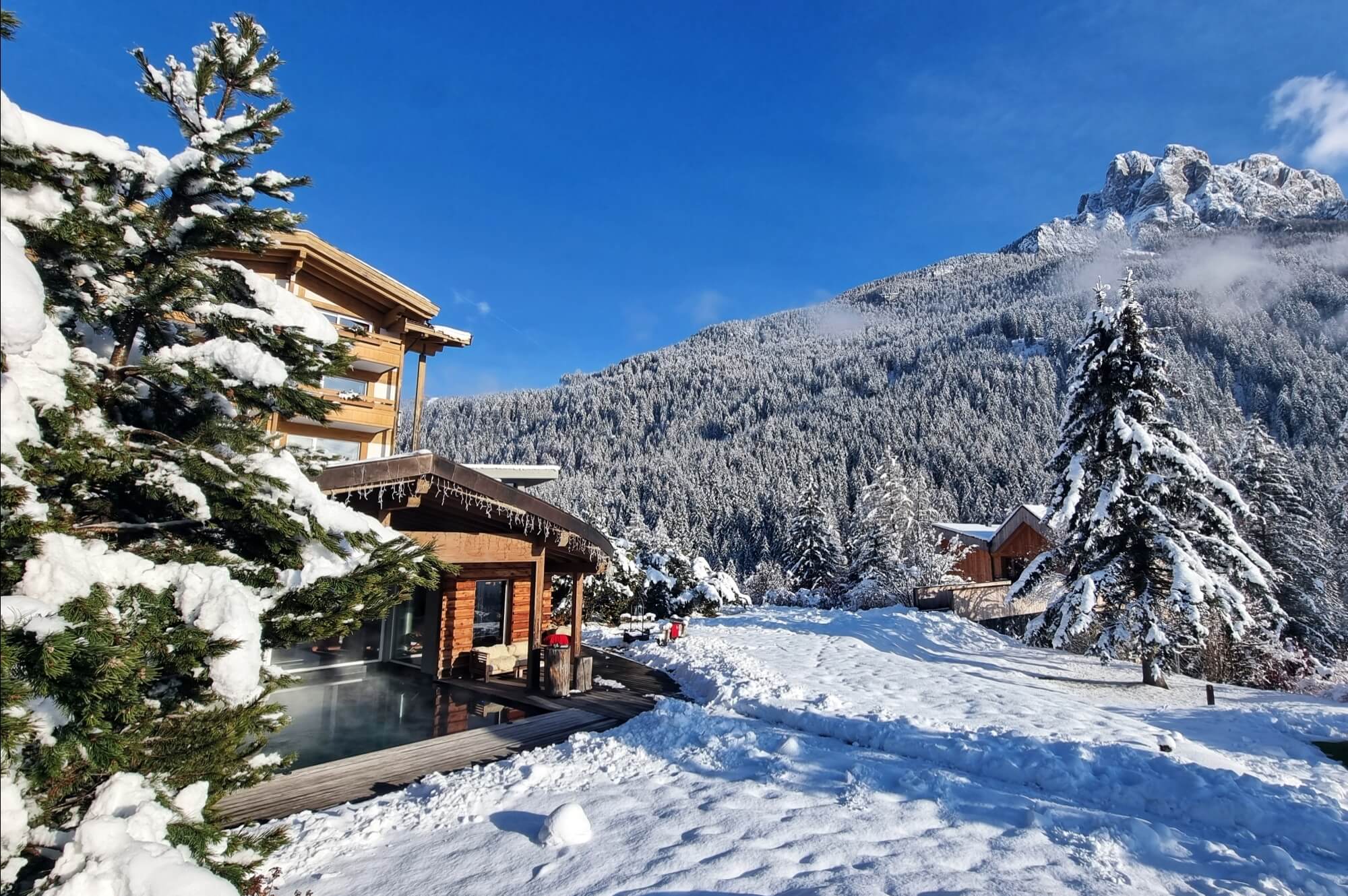Una cabina di legno in un paesaggio invernale sereno, circondata da montagne e alberi sempreverdi coperti di neve.