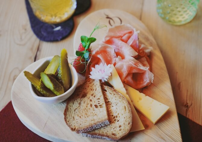Tagliere in legno con pane, formaggio, prosciutto e cetrioli, posizionato su un tavolo con altri oggetti sullo sfondo.