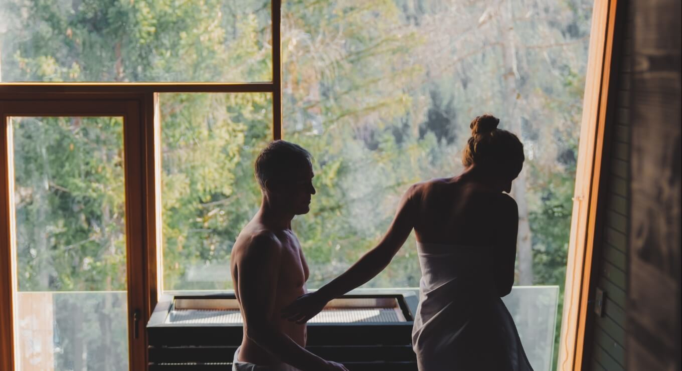 Uomo e donna in sauna, lui senza maglietta e lei con un asciugamano, entrambi in controluce davanti a una finestra.