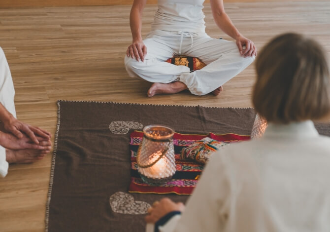 Tre persone in meditazione su un pavimento di legno, vestite di bianco, con candele e un tappeto colorato.