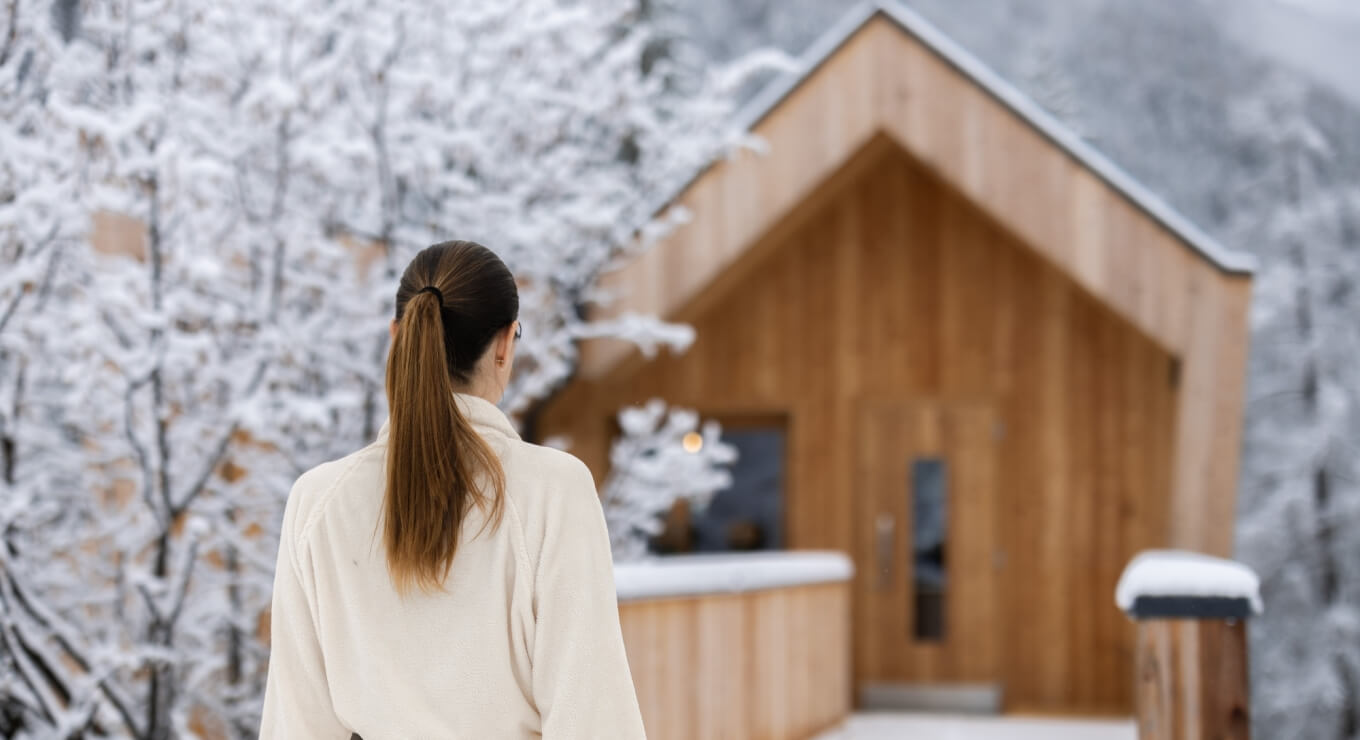 Donna in cappotto bianco davanti a una casa di legno moderna, con neve, capelli castani raccolti in una coda di cavallo.
