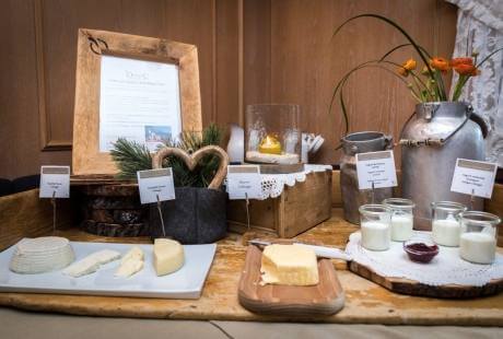 Un tavolo di legno con formaggi, latte in barattoli, fiori arancioni, segni bianchi e un telaio di legno con rami di pino.