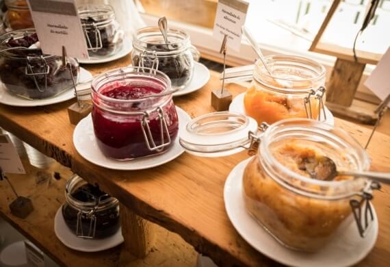 Vetrina di barattoli di marmellata su piatti bianchi, con cucchiai e segnalini, illuminata da luce naturale.