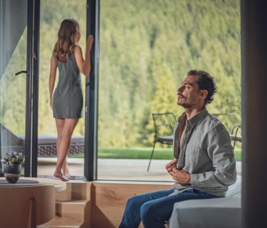 Una donna in abito grigio sta fuori da una finestra mentre un uomo in maglietta grigia la guarda seduto su un divano.