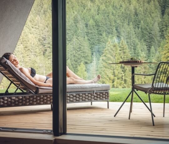Una donna rilassata su una sdraio, con vista su una foresta attraverso una parete di vetro, trasmette serenità.