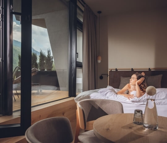 Una donna sdraiata su un letto in una stanza accogliente, persa nei suoi pensieri, con vista su alberi e montagne.