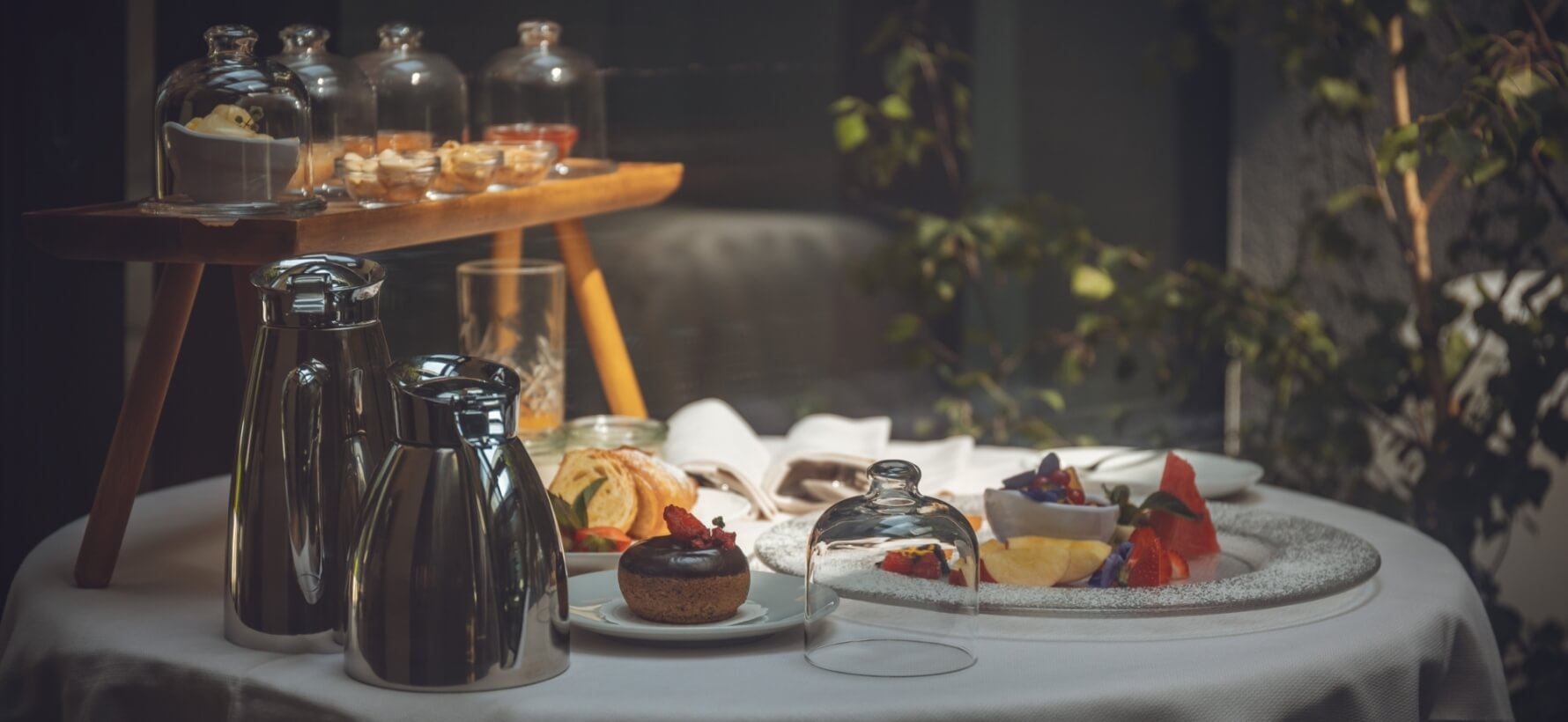 Tavolo con tovaglia bianca, caraffe d'argento, piatti di dolci e frutta, pianta e carrello in un ambiente poco illuminato.