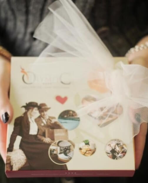 Una donna con un vestito scuro tiene un regalo con un collage vintage e un fiocco bianco, mani con unghie nere.