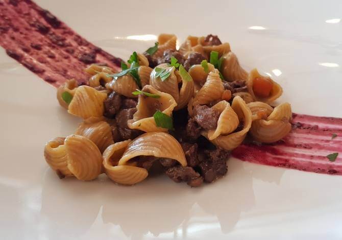Piatto di pasta con carne e erbe, con salsa rossa sullo sfondo, riflesso di una finestra sulla superficie bianca.