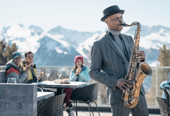 Uomo in abito grigio suona il sassofono con montagne innevate sullo sfondo.