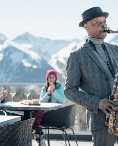 Uomo in abito grigio suona il sassofono con montagne innevate sullo sfondo.