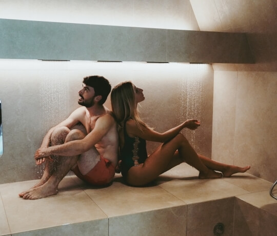 Uomo e donna seduti in una doccia, schiena contro schiena, in un ambiente intimo e rilassante con piastrelle beige.