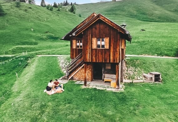 Una piccola casa di legno in una valle verde, con una coppia seduta su una coperta e un cielo sereno sopra di loro.