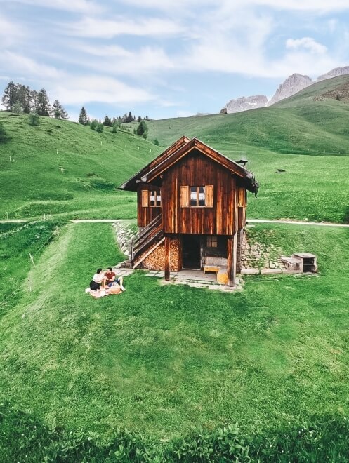 Una casa di legno in un campo verde, con due persone sedute su un prato, circondate da colline e un cielo blu.
