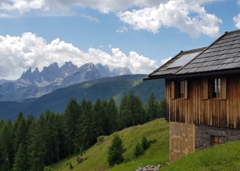 Casa in legno con pannelli solari, circondata da alberi e montagne, sotto un cielo blu con nuvole bianche.