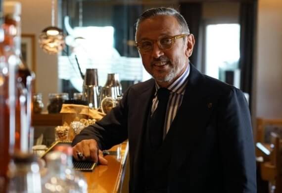 Uomo in giacca nera al bar, con occhiali e anello, circondato da bottiglie e bicchieri sfocati sullo sfondo.