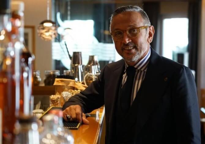 Uomo in abito nero dietro un bar, con una mano su uno strumento, circondato da bottiglie e oggetti da mixology.