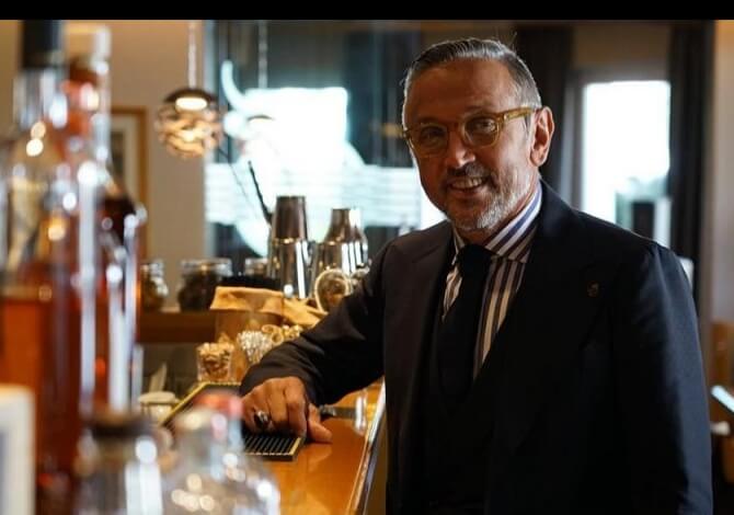 Uomo in abito elegante dietro un bar, con occhiali e penna su un taccuino, in un ambiente sofisticato e luminoso.