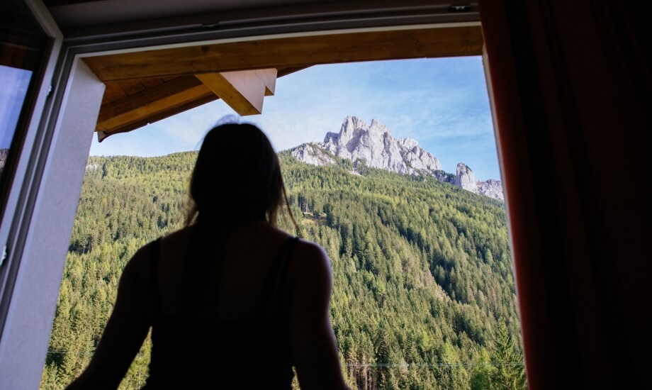 Una donna in silhouette guarda un paesaggio montano attraverso una finestra aperta, circondata da un'atmosfera serena.