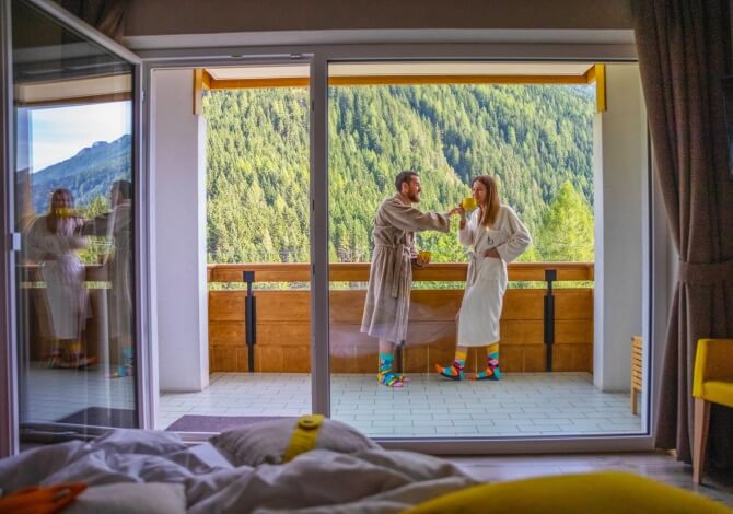 Una coppia in accappatoi bianchi e calzini colorati su un balcone con vista su foreste e montagne, in un'atmosfera serena.