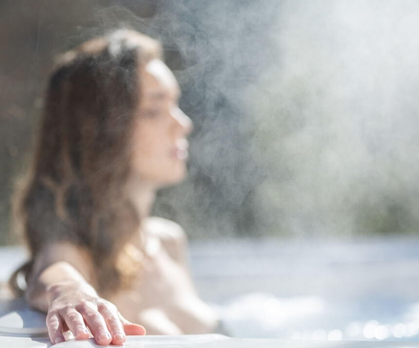 Una donna rilassata in una vasca idromassaggio, con la mano destra appoggiata sul bordo, circondata da un ambiente naturale sfocato.