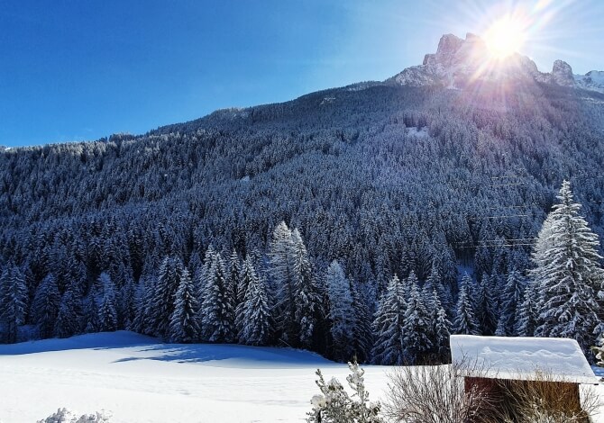 Scena invernale serena con montagne innevate, alberi, cielo blu e un piccolo edificio coperto di neve.