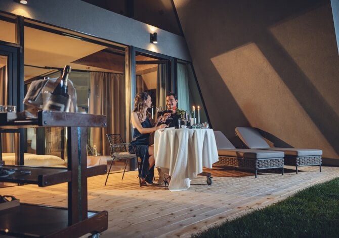 Coppia a cena romantica su un deck moderno, tavolo con fiori rossi, candele e atmosfera calda.