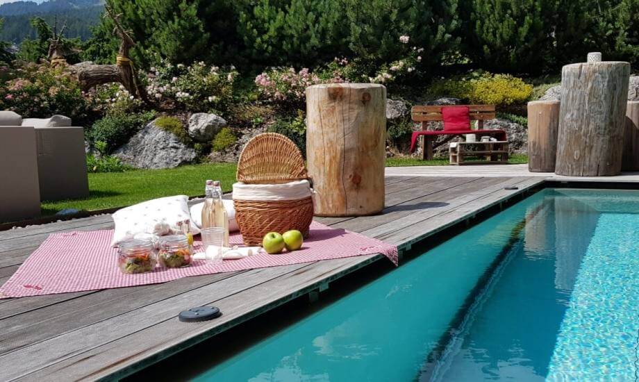 Immagine di un'area picnic serena con piscina, tavolo a quadretti rossi e bianchi, cibo e natura lussureggiante.