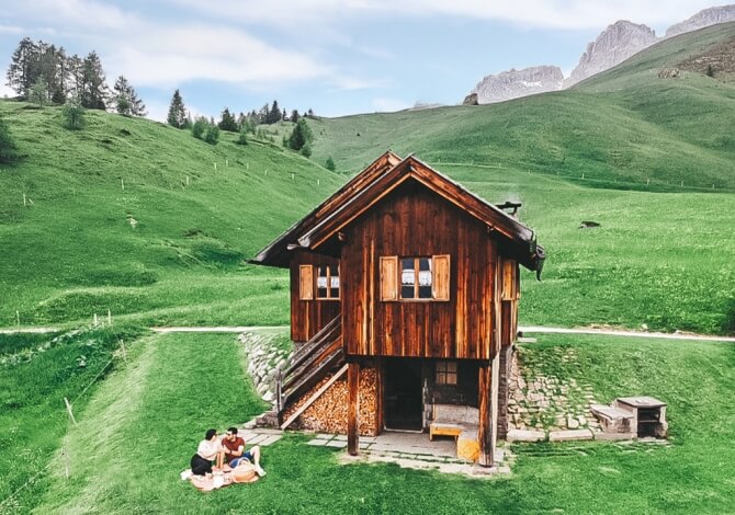 Una piccola casa di legno circondata da un paesaggio verde, con due persone sedute su una coperta e montagne in lontananza.