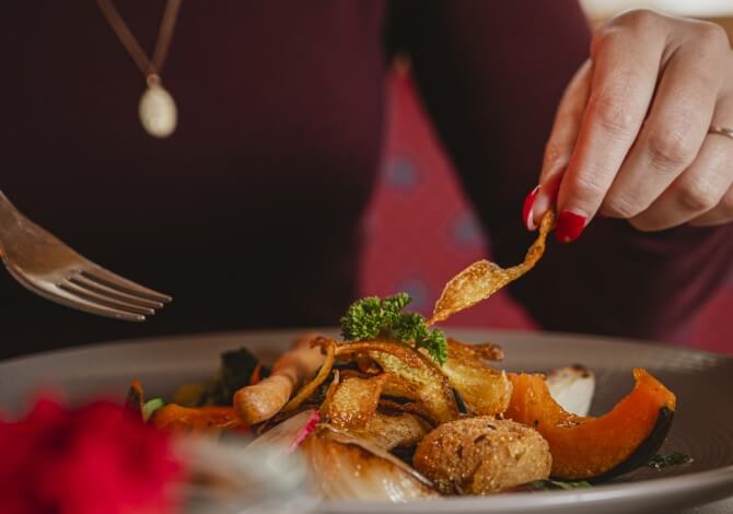 Donna con un top rosso scuro, tiene un pezzo di cibo fritto e una forchetta sopra un piatto di cibo misto.