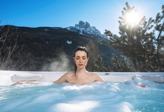Una donna rilassata in una vasca idromassaggio all'aperto, circondata da montagne maestose e vegetazione lussureggiante.