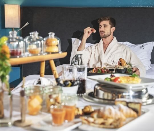Uomo in pigiama bianco seduto a letto, circondato da una ricca colazione su un tavolo bianco. Atmosfera lussuosa.