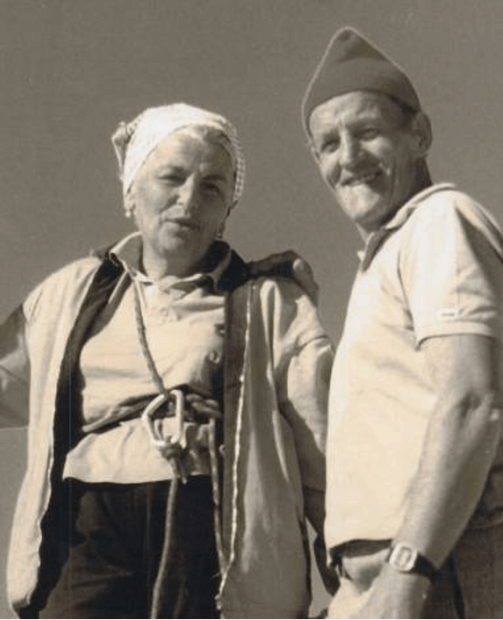 Due persone in primo piano: una donna con foulard e giacca, un uomo sorridente con cappellino e camicia chiara.