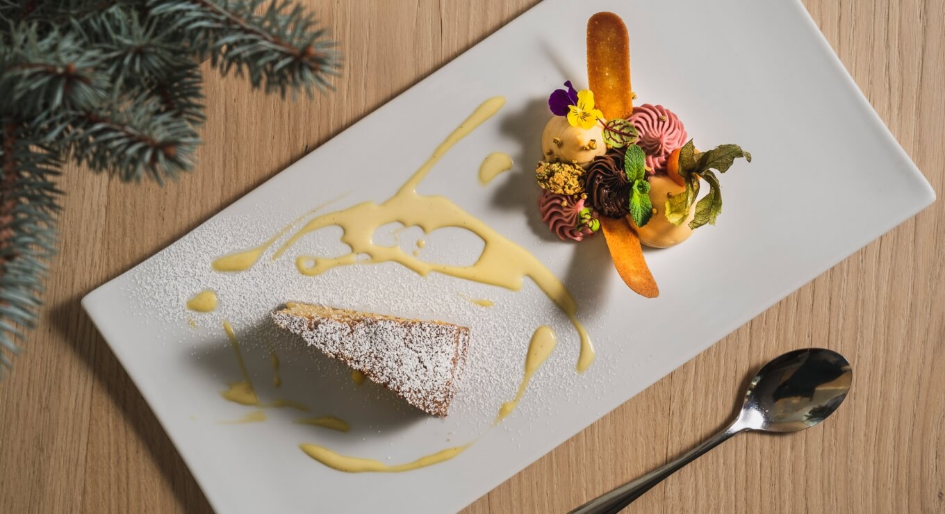 Piatto bianco con torta quadrata spolverata di zucchero a velo e dessert colorato con fiori, foglie e salse.