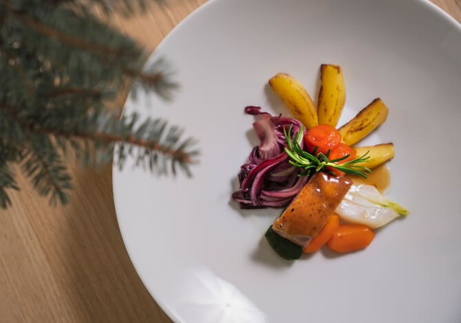 Piatto bianco con verdure colorate, pesce o platano, rametto di rosmarino e un ramo di abete natalizio.