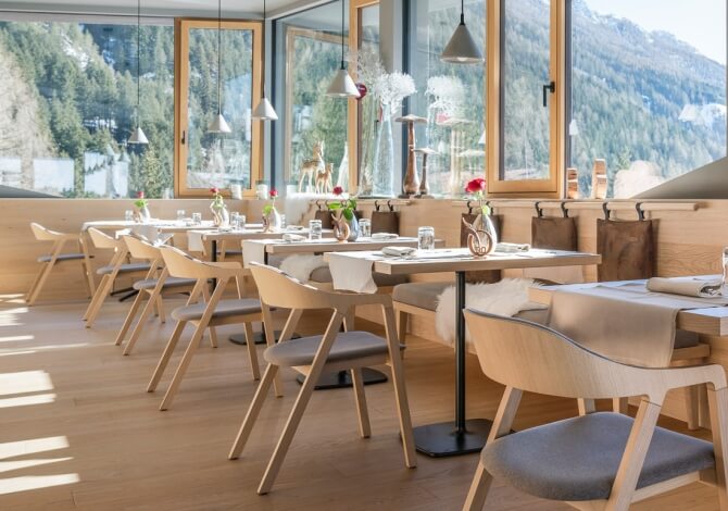 Area ristorante moderna con tavoli bianchi, fiori rossi e vista montuosa attraverso grandi finestre.