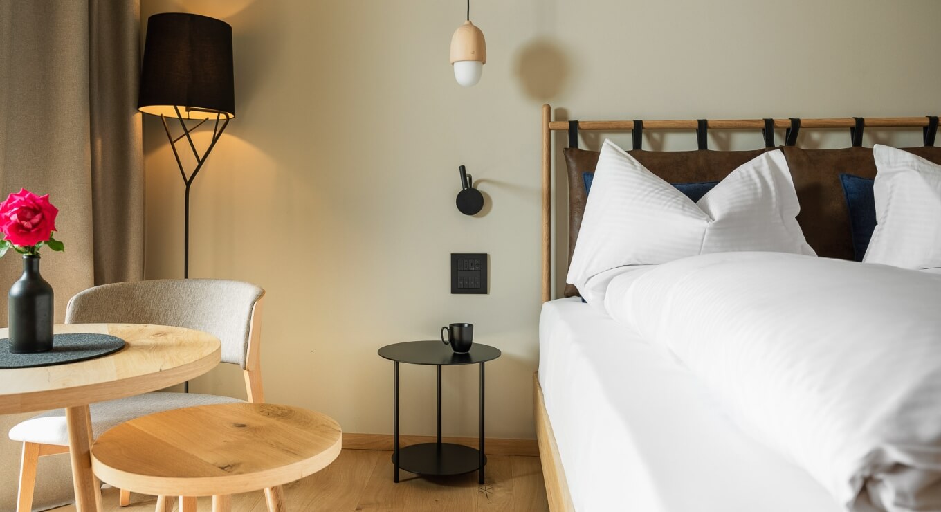 Camera elegante e serena con letto bianco, tavolo rotondo nero, fiori rosa e luce soffusa. Atmosfera di calma e raffinatezza.