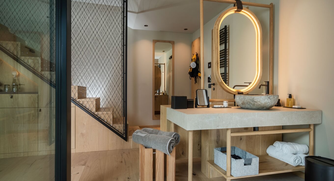 Bagno moderno con vanity in legno chiaro, lavabo in pietra, specchi e sgabelli, pavimento in legno chiaro e scala sullo sfondo.