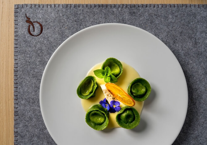 Piatto bianco con salsa gialla, guarnito con verdure verdi, menta, fetta d'arancia e fiore viola su un sottopiatto grigio.