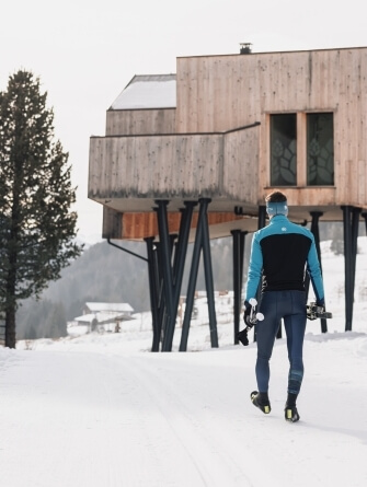 Uomo in rollerblade sulla neve, con arco, vicino a una casa di legno su stilts, in un paesaggio invernale.