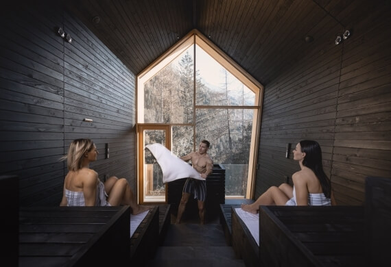Due donne in sauna e un uomo con asciugamano, vista su alberi.