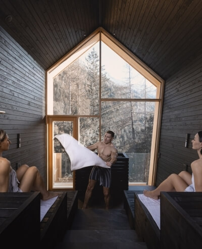 Spa interior with wooden benches, two women, and a man. Interno di una spa con panche di legno, due donne e un uomo.