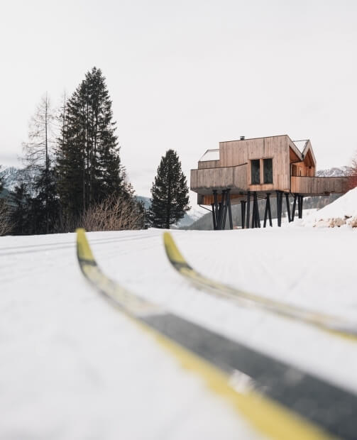 Casa moderna in legno elevata su pali neri in un paesaggio innevato, con sci gialli e neri in primo piano.
