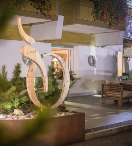 Scultura in legno chiaro con forme curve, piante in un vaso, panchine e ingresso di un edificio in un ambiente sereno.