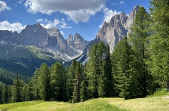 Vista di picchi rocciosi con neve, valle verde con alberi sempreverdi e cielo azzurro con nuvole bianche.