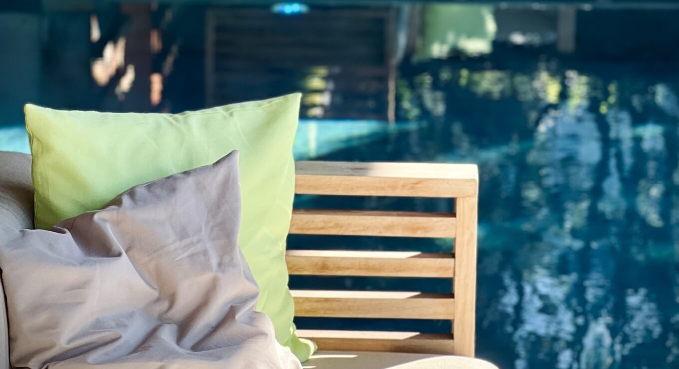 Area esterna serena con una sedia di legno e tre cuscini colorati: verde lime, grigio-marrone e beige.