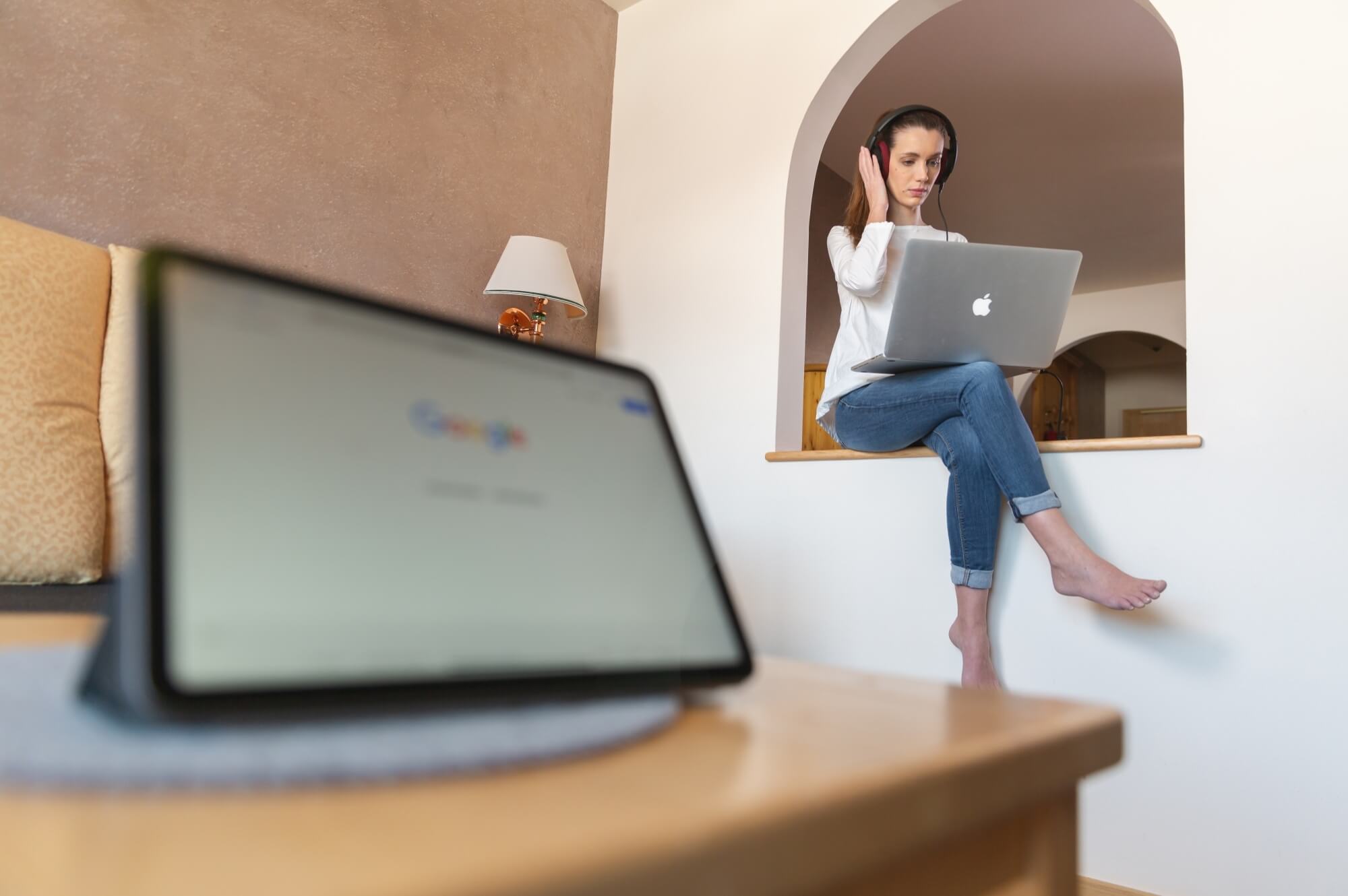 Donna seduta su un ledge con laptop, indossando cuffie, in una stanza con letto e lampada sul comodino.